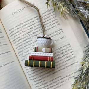 Könnte beinhalten: Handgefertigtes Lesezeichen mit einem Stapel Miniaturbücher und einem Teetassen-Anhänger. Die Bücher sind in Rot, Grün und Creme gehalten, mit einer Juteschnur zum Aufhängen. Das Lesezeichen liegt auf den offenen Seiten eines Buches.
