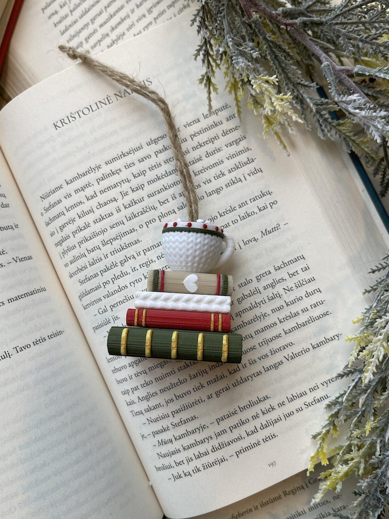 Puede incluir: Marcap&aacute;ginas artesanal con una pila de libros en miniatura y un dije de taza de t&eacute;. Los libros son de color rojo, verde y crema, con una cuerda de yute para colgar. El marcap&aacute;ginas descansa sobre las p&aacute;ginas abiertas de un libro.