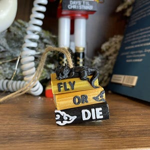 May include: A black dragon ornament rests atop a stack of books with the words "FLY OR DIE" in white. The books are yellow and black. A brown string is attached for hanging. A Christmas nutcracker and greenery are in the background.