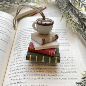 Puede incluir: Un marcap&aacute;ginas caprichoso con una peque&ntilde;a taza de caf&eacute; blanca con una bebida marr&oacute;n y malvaviscos, encima de una pila de tres libros coloridos. El marcap&aacute;ginas est&aacute; hecho de arcilla polim&eacute;rica y tiene una cuerda de yute para colgar.