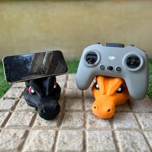 Może przedstawiać: Dwa uchwyty w kształcie smoka, jeden czarny na telefon, drugi pomarańczowy na szary kontroler. Oba stoją na kamiennej powierzchni, prezentując oryginalny design akcesoriów technologicznych.