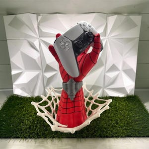 Pode incluir: Uma escultura de mão vermelha e preta com um padrão de teia de aranha segurando um controle de jogo sem fio branco e preto. A mão apresenta um punho cinza e emerge de uma estrutura de teia tridimensional branca na base.