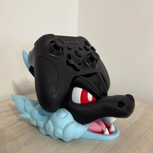 以下が含まれることがあります： 黒いゲームコントローラーが、水色のドラゴンのテーマのスタンドに置かれています。スタンドは、赤い目、白い歯、ピンクの舌を持つドラゴンの頭が特徴です。コントローラーは黒色です。