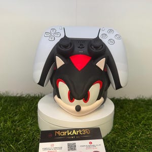 Può includere: Una figura nera, rossa e bianca di Shadow the Hedgehog con un controller PlayStation 5. La figura è su un piedistallo bianco, con un biglietto da visita davanti. La carta ha il testo "MarkArt3D PRINT 3D".