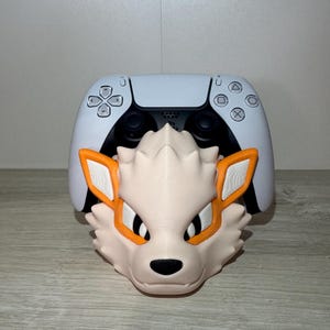 May include: A light tan and orange Arcanine-themed controller holder with a white PlayStation 5 controller in the background. The Arcanine head has black eyes and a black nose. The controller has the standard PlayStation button layout.
