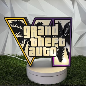 Pode incluir: Um letreiro luminoso em forma de V com o logótipo Grand Theft Auto em letras brancas sobre um fundo preto com silhuetas de palmeiras. O letreiro tem um rebordo amarelo e roxo e está montado numa base circular branca com um cabo.