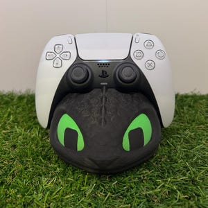 Może przedstawiać: Biało-czarny kontroler PlayStation 5 spoczywa na czarnej głowie smoka z zielonymi oczami. Głowa smoka to podstawka pod kontroler, ustawiona na zielonej sztucznej trawie.