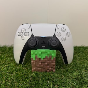 Może przedstawiać: Biało-czarny kontroler PlayStation 5 spoczywa na bloku w stylu Minecraft. Kontroler ma elegancki design z różnymi symbolami przycisków. Blok jest brązowy i zielony, przypominający pikselowy blok trawy i ziemi.
