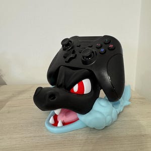 以下が含まれることがあります： 黒いゲームコントローラーが、黒と青のドラゴンをテーマにしたスタンドに置かれています。ドラゴンは赤い目、白い歯、水色の雲のようなベースをしています。コントローラーには色の付いたボタンがあります。