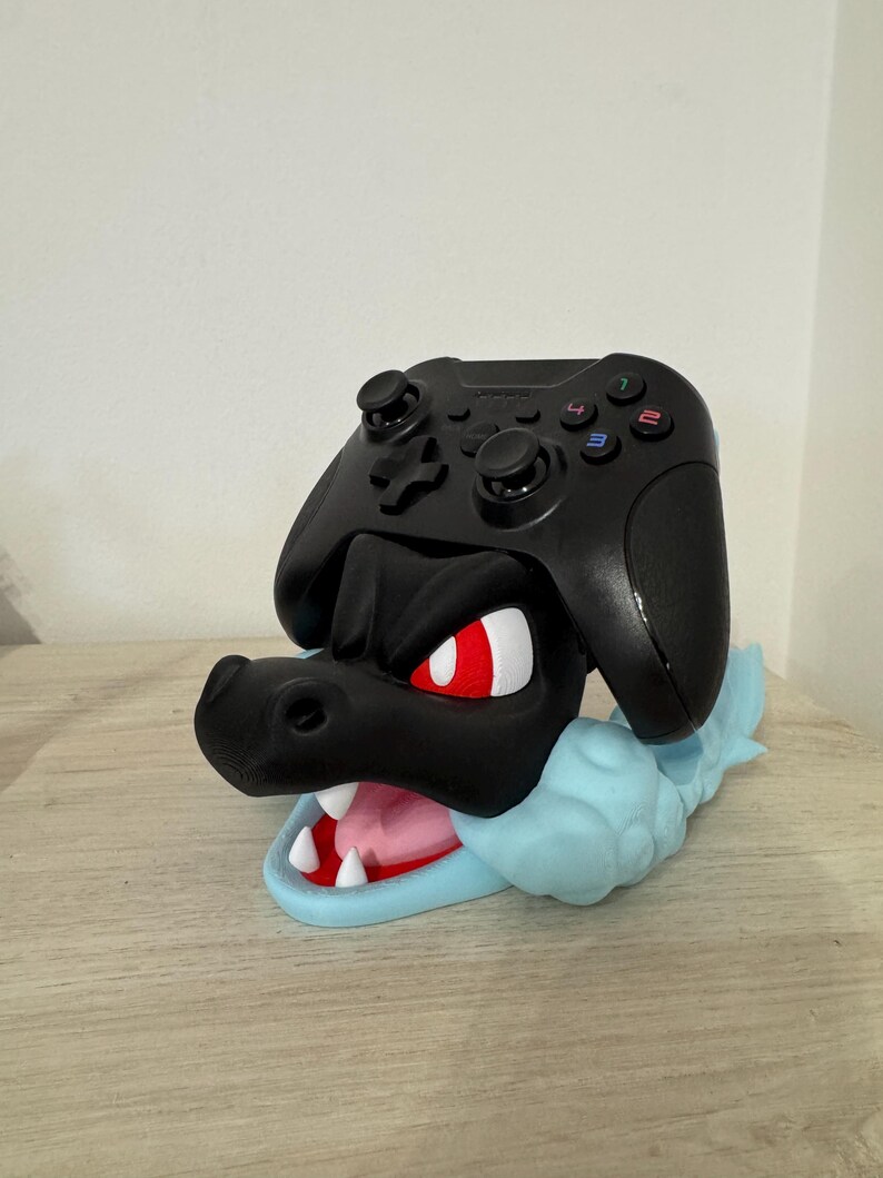 以下が含まれることがあります： 黒いゲームコントローラーが、黒と青のドラゴンをテーマにしたスタンドに置かれています。ドラゴンは赤い目、白い歯、水色の雲のようなベースをしています。コントローラーには色の付いたボタンがあります。