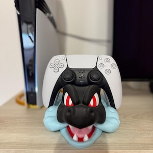 以下が含まれることがあります： 赤目と白い歯を持つ黒と青のドラゴンをテーマにしたコントローラーのスタンドで、白と黒のPlayStation 5コントローラーを持っています。スタンドは木製の表面にあります。