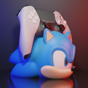 May include: A blue Sonic the Hedgehog controller holder with a grey and white game controller on top. The holder is shaped like Sonic's head and body. The background has a gradient of red and blue.