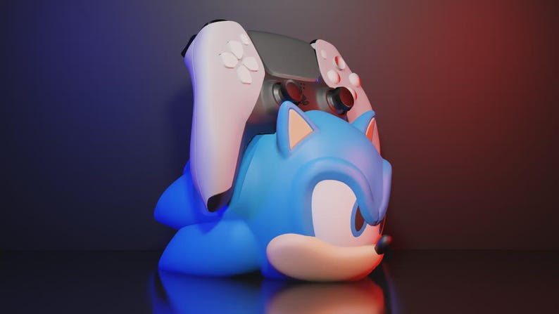 May include: A blue Sonic the Hedgehog controller holder with a grey and white game controller on top. The holder is shaped like Sonic's head and body. The background has a gradient of red and blue.