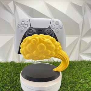 Op de afbeelding: Een wit en zwart videogame-controller rust op een gele, wolk-vormige standaard. De standaard heeft een getextureerd, wervelend wolkenontwerp dat uitsteekt vanaf een ronde zwarte basis, gemonteerd op een witte cilindrische voet.