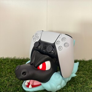以下が含まれることがあります： 黒と青のドラゴンをテーマにしたコントローラーホルダー。白いPlayStation 5コントローラーが上に置かれています。ドラゴンは赤い目とピンクの口をしています。ホルダーは緑色の表面にあります。