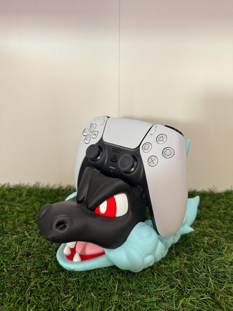 以下が含まれることがあります： 黒と青のドラゴンをテーマにしたコントローラーホルダー。白いPlayStation 5コントローラーが上に置かれています。ドラゴンは赤い目とピンクの口をしています。ホルダーは緑色の表面にあります。