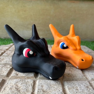 Pode incluir: Duas figuras de cabe&ccedil;a de drag&atilde;o, uma preta e outra laranja, lado a lado. O drag&atilde;o preto tem olhos vermelhos e brancos, enquanto o drag&atilde;o laranja tem olhos azuis. Ambos t&ecirc;m orelhas pontudas e uma textura detalhada.