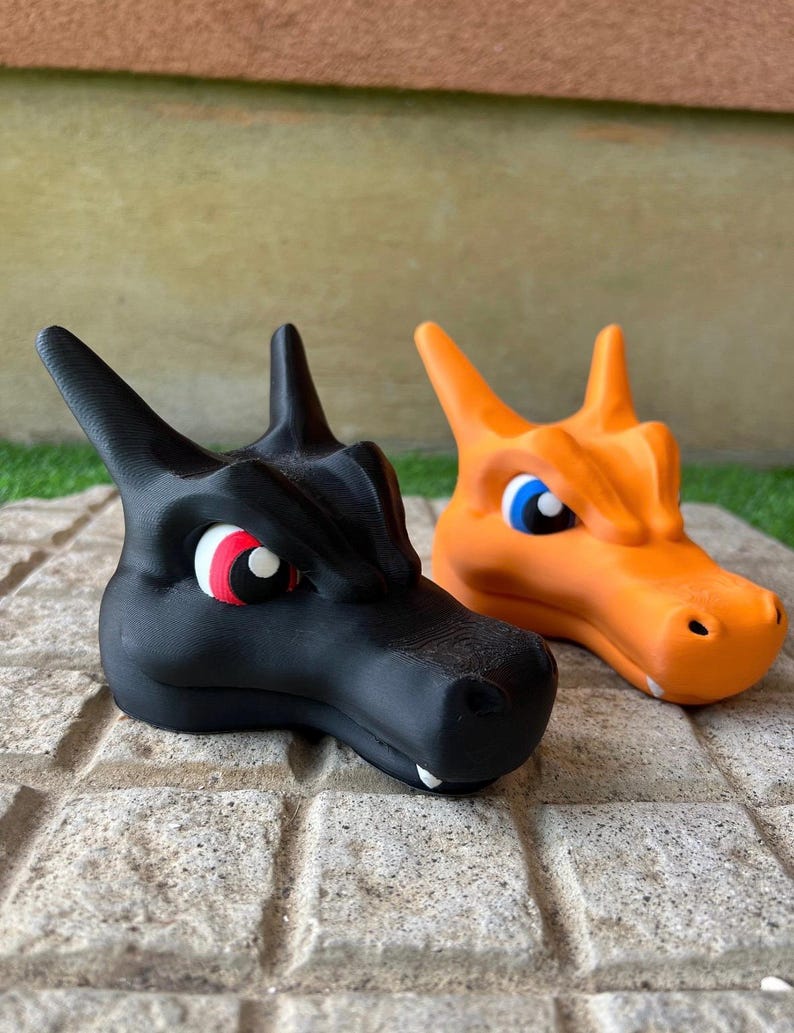 Pode incluir: Duas figuras de cabe&ccedil;a de drag&atilde;o, uma preta e outra laranja, lado a lado. O drag&atilde;o preto tem olhos vermelhos e brancos, enquanto o drag&atilde;o laranja tem olhos azuis. Ambos t&ecirc;m orelhas pontudas e uma textura detalhada.