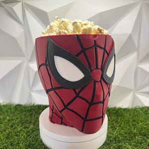 Può includere: Una ciotola rossa e nera a tema Spider-Man riempita di popcorn. La ciotola ha la forma della testa del personaggio, con grandi occhi bianchi dai contorni neri e un motivo a ragnatela nera sulla superficie rossa. Poggia su una base circolare.