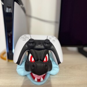 以下が含まれることがあります： 黒と赤のドラゴンをテーマにしたコントローラーホルダー。白と黒のゲームコントローラーが上に置かれています。ドラゴンは赤い目を持ち、青いベースが付いています。ホルダーはコントローラーを支えるように設計されています。