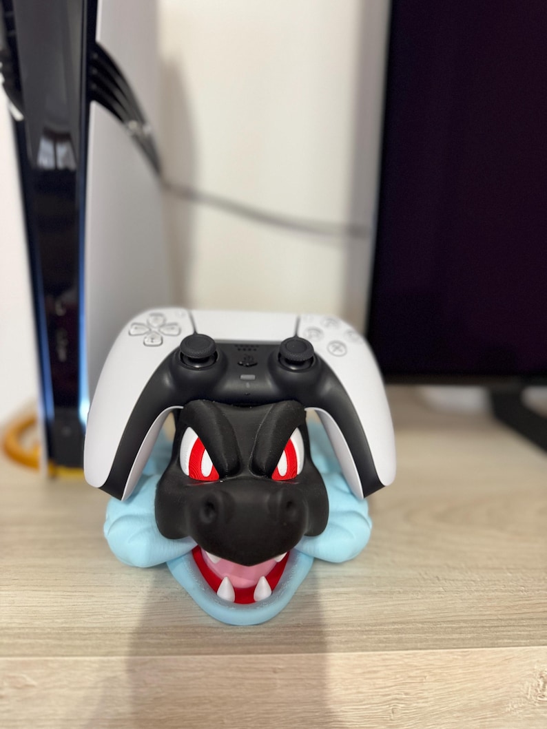 以下が含まれることがあります： 黒と赤のドラゴンをテーマにしたコントローラーホルダー。白と黒のゲームコントローラーが上に置かれています。ドラゴンは赤い目を持ち、青いベースが付いています。ホルダーはコントローラーを支えるように設計されています。