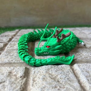 Könnte beinhalten: Grüne Drachenfigur mit roten Augen und braunen Geweihen, auf einer Steinoberfläche aufgerollt. Der Drachenkörper ist segmentiert. Der Kopf hat spitze Zähne und Schnurrhaare. Ungefähr 15 cm lang.