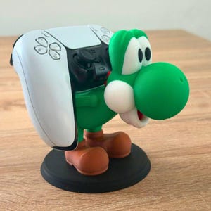 Può includere: Un supporto per controller a tema Yoshi verde che tiene in mano un controller di gioco bianco e nero. Il supporto presenta una figura di Yoshi verde con dettagli bianchi, scarpe marroni e una base nera. Il controller è bianco con pulsanti neri.