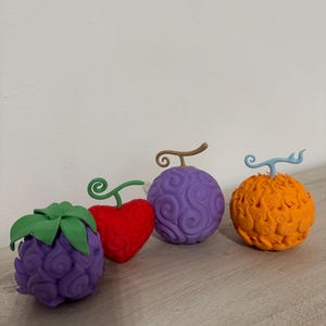 May include: Four colorful, stylized fruit sculptures. A purple, berry-like fruit with green leaves, a red heart-shaped fruit, a purple sphere with a swirl pattern, and an orange sphere with a curly pattern.
