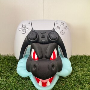 以下が含まれることがあります： 白のPlayStation 5コントローラーを保持するように設計された、黒と赤のドラゴンをテーマにしたコントローラーホルダー。ライトブルーのアクセント付き。ホルダーは緑色の人工芝の上に配置されています。