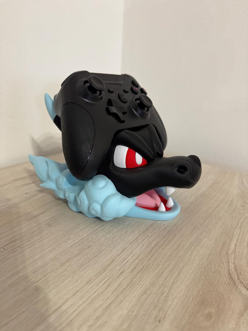 以下が含まれることがあります： 黒いゲームコントローラーが、水色のドラゴンのテーマのスタンドに置かれています。スタンドは、赤い目、白い歯、ピンクの舌を持つドラゴンの頭が特徴です。コントローラーは黒色です。