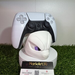 May include: A white and black PlayStation 5 controller rests atop a white 3D-printed sculpture of a character's head with purple eyes. The sculpture sits on a white pedestal. A business card with the text "MarkArt39 PRINT 3D" is in front.