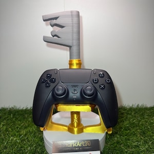 Könnte beinhalten: Ein schwarzer Videospiel-Controller ruht auf einem gold-weißen Ständer, der von einem grau geformten Schlüsseldesign gekrönt wird. Der Ständer hat einen goldenen Sockel und eine weiße Plattform. Eine Visitenkarte mit dem Text "MarkArt39" befindet sich am Boden.