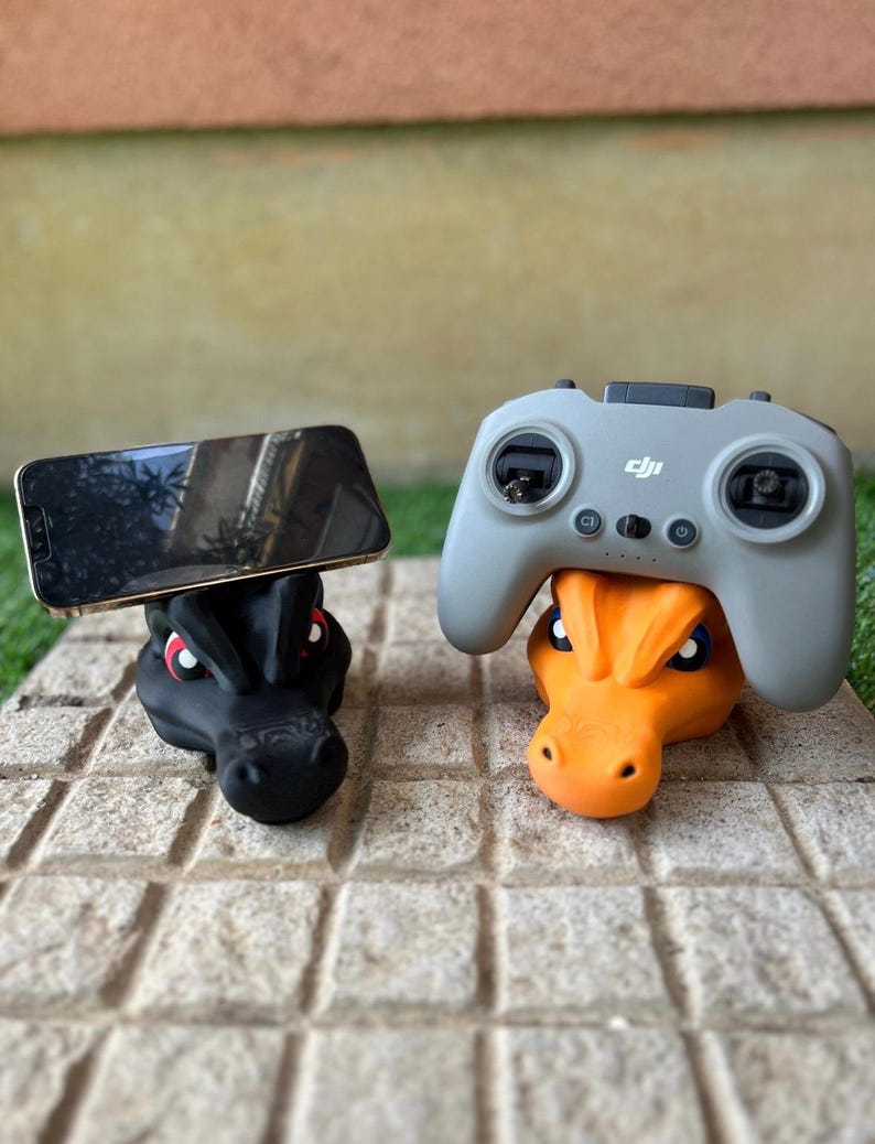 Pode incluir: Dois suportes em forma de drag&atilde;o, um preto segurando um telefone, outro laranja suportando um controle cinza. Ambos est&atilde;o em uma superf&iacute;cie de pedra, mostrando um design original.