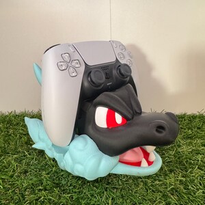 以下が含まれることがあります： 黒と水色のドラゴンをテーマにしたコントローラーホルダー。グレーのコントローラーが上に置かれています。ドラゴンは赤い目と白い歯をしています。ホルダーは緑色の表面にあります。