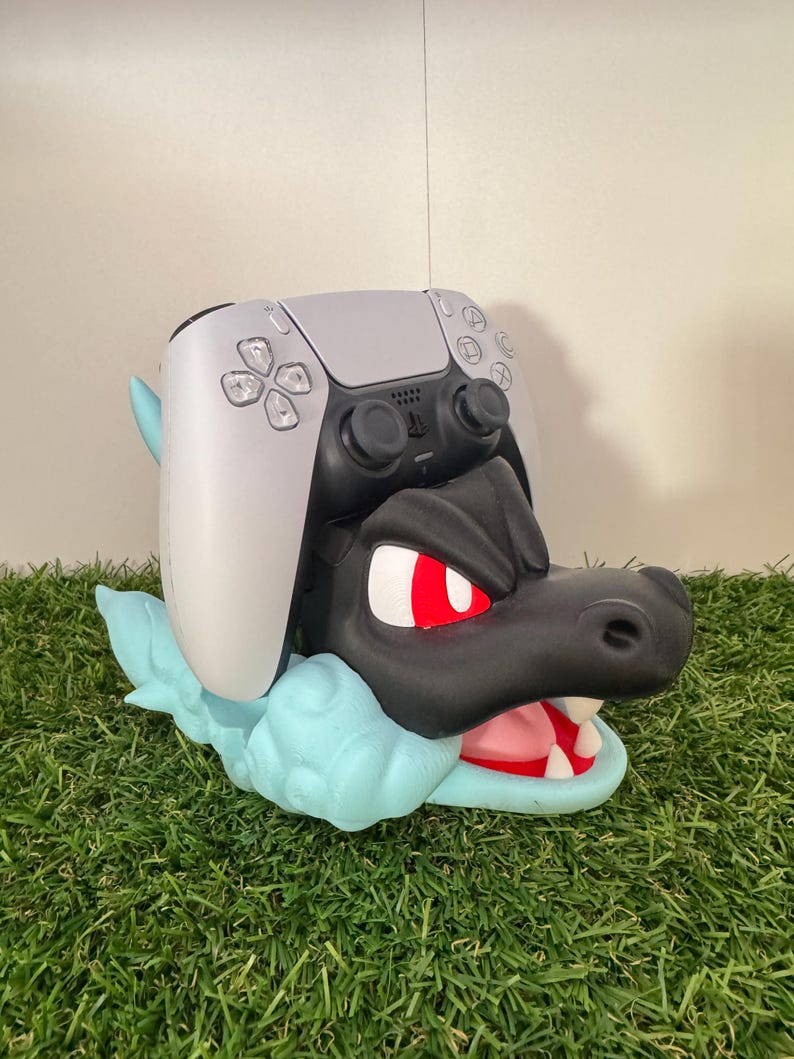 以下が含まれることがあります： 黒と水色のドラゴンをテーマにしたコントローラーホルダー。グレーのコントローラーが上に置かれています。ドラゴンは赤い目と白い歯をしています。ホルダーは緑色の表面にあります。