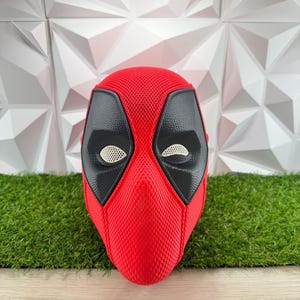 Könnte beinhalten: Eine rot-schwarze Vollkopfmaske mit einer strukturierten Stoffoberfläche. Die Maske hat große, schwarze Augenpartien mit weißen Netzgläsern, die das Aussehen der Figur Deadpool nachahmen. Das Design weist eine markante Naht in der Mitte auf.
