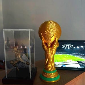 May include: A gold-colored FIFA World Cup trophy replica with a green base, positioned next to a clear acrylic display case containing a small metallic figurine of a soccer player in a dynamic kicking pose on a black stand.
