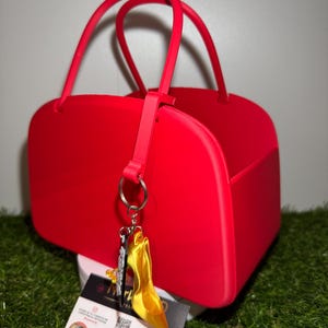 Puede incluir: Un bolso rojo brillante con dos asas curvas y forma rectangular. Un charm de zapato de tacón alto amarillo cuelga del asa. El bolso está sobre una superficie blanca con una tarjeta de visita y un fondo verde y herboso.