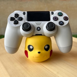 May include: A white PlayStation 4 controller rests atop a yellow Pikachu figure. The controller features black buttons and directional pad, with colored action buttons. The Pikachu figure has black eyes and red cheeks, with a smiling expression.