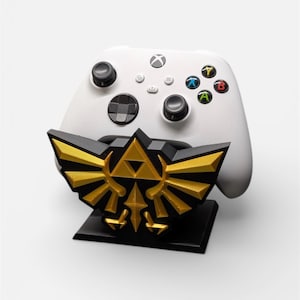 Op de afbeelding: Een witte Xbox-controller rust op een zwart en gouden decoratieve standaard. De standaard heeft een gouden embleem met een zwarte basis. De controller heeft gekleurde knoppen en zwarte joysticks.