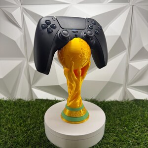Könnte beinhalten: Ein schwarzer Videospiel-Controller auf einem goldfarbenen Trophäenständer. Die Trophäe hat eine kugelförmige Spitze und einen Sockel mit dem grün geprägten Text FIFA WORLD CUP. Der Ständer ist so konzipiert, dass er den Controller sicher hält.