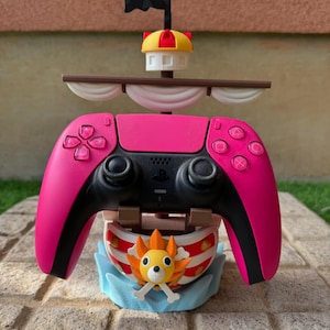May include: A pink and black video game controller with a ship-themed stand. The stand features a cartoon ship with a black flag, a yellow and red mast, and a cartoon character with crossbones. The controller has pink buttons.