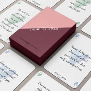 May include: A card game set featuring a rectangular box with a diagonal split of light pink and dark maroon. The box is labeled DRAW US CLOSER. Surrounding the box are several white playing cards with black text, light blue and green heart icons, and the brand name DRAW US CLOSER printed at the bottom of each card.