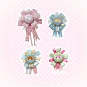 Peut inclure: Quatre badges décoratifs en forme de rosette avec un visage d'animal de dessin animé au centre. Les badges présentent diverses couleurs, dont le rose, le bleu et le vert, avec des rubans et des accents de dentelle. Le fond est rose clair avec des pois blancs.