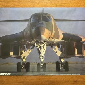 May include: A B1 Bomber aircraft poster. The aircraft is on the ground, viewed head-on, with its landing gear deployed. The plane has a camouflage pattern of brown, green, and tan. The text "B1 Bomber" is at the bottom.
