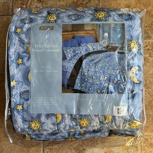 Puede incluir: Un edredón de franela Full/Queen en un paquete de plástico. El edredón tiene un fondo azul con soles, lunas y estrellas amarillas. El paquete tiene una foto del edredón en una cama. El edredón mide 228 cm x 228 cm.