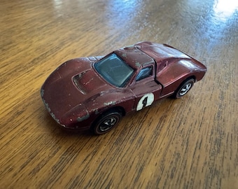 May include: A vintage burgundy die-cast toy car with black wheels. The car has a white number 1 on the side. The paint shows signs of wear, indicating its age. The car is a classic collectible.