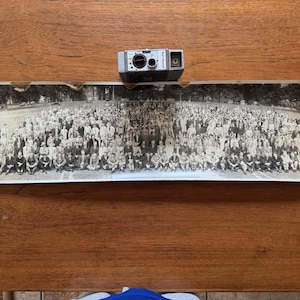 May include: A small, silver vintage camera sits on a long, rectangular tray. The tray features a black and white photograph of a large group of people. The tray is on a wooden surface.