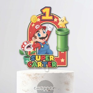 Peut inclure: Un cake topper color&eacute; sur le th&egrave;me de Super Mario Bros. avec Mario, un tuyau vert et le chiffre 1. Le texte indique "Super Carter". Le g&acirc;teau est blanc avec des d&eacute;tails dor&eacute;s, pr&eacute;sent&eacute; sur un pr&eacute;sentoir &agrave; g&acirc;teau en bois.