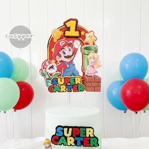 Peut inclure: Un g&acirc;teau d'anniversaire sur le th&egrave;me de Super Mario, avec un d&eacute;grad&eacute; vert et blanc. Le g&acirc;teau est d&eacute;cor&eacute; d'un topper Mario, d'une figurine de la princesse Peach et du texte "SUPER CARTER". Des ballons rouges, bleus et verts compl&egrave;tent l'ensemble festif.